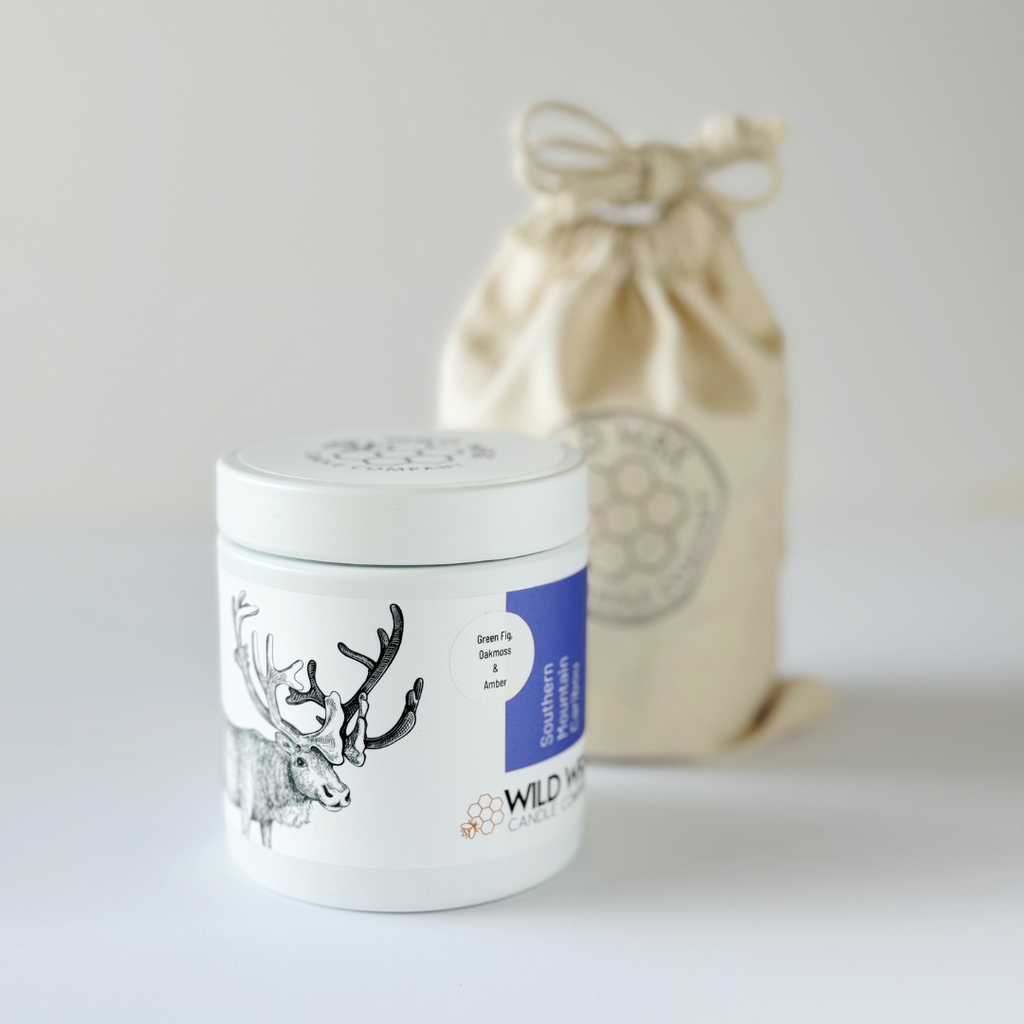Minimal white candle container with lid featuring caribou illustration in front of linen drawstring gift bag 
